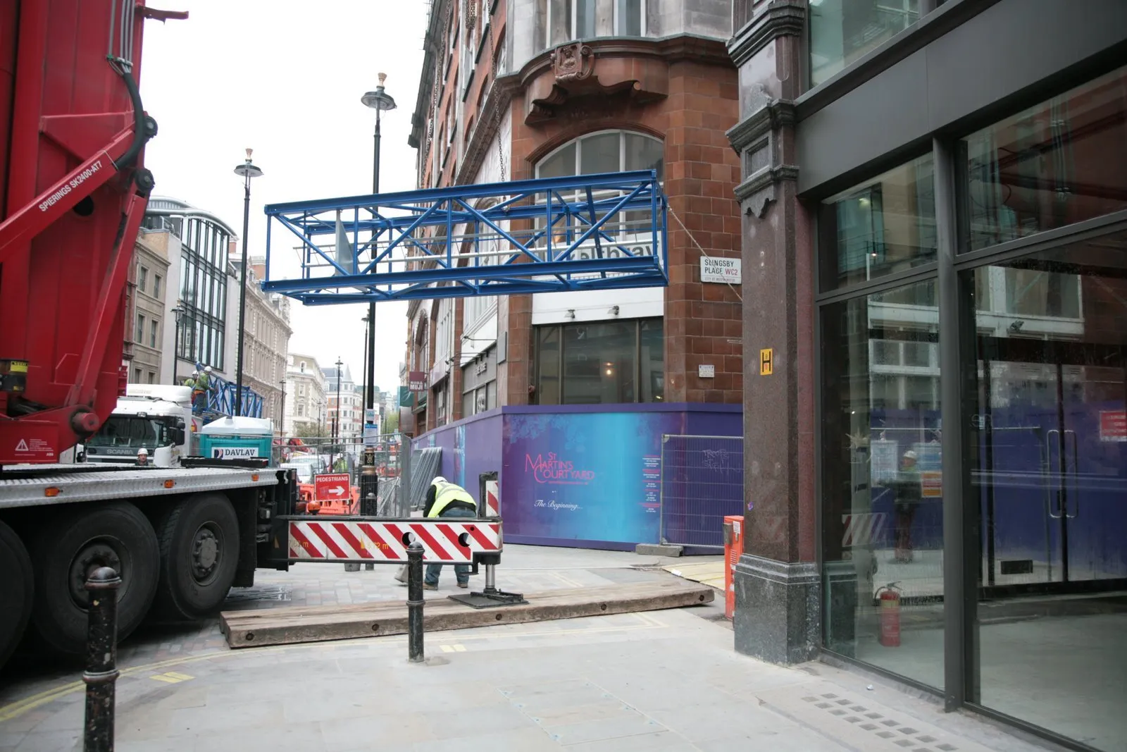 Mobile crane lifting a tower-crane section on a Covent Garden street beside site hoarding at Slingsby Place