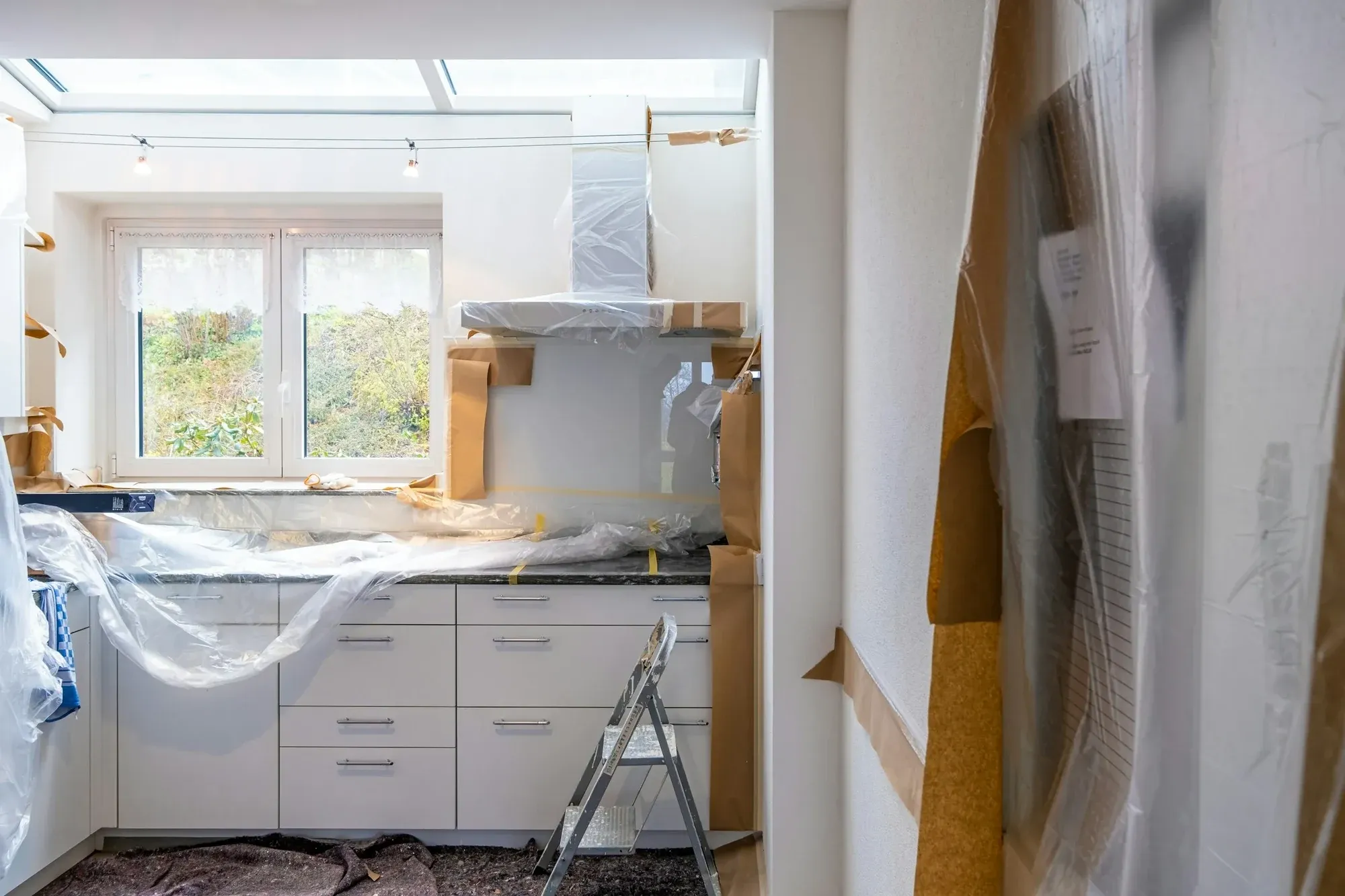 Kitchen mid-renovation with units and worktops wrapped in plastic sheeting and a stepladder on the floor